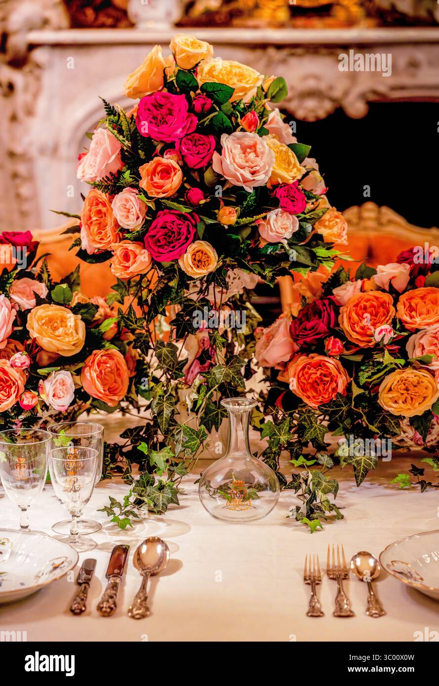 Sfarzosa rosa al centro di rose di corallo, rosa e pesca intrecciata con l'edera che adorna un tavolo da banchetto elegantemente allestito sotto una calda luce di candela Foto Stock
