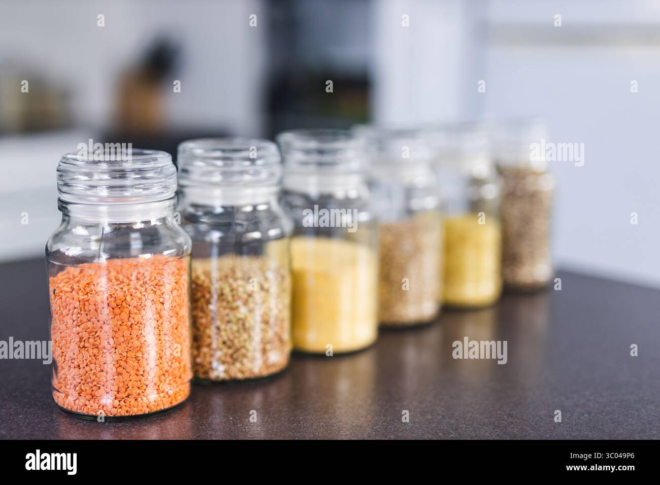 vasi di pantry trasparenti con diversi tipi di cereali e legumi, tra cui quinoa lenticchie grano saraceno e orzo sul banco con bokeh da cucina, semplice ing Foto Stock vasi di pantry trasparenti con diversi tipi di cereali e legumi, tra cui quinoa lenticchie grano saraceno e orzo sul banco con bokeh da cucina, semplice ing Foto Stock