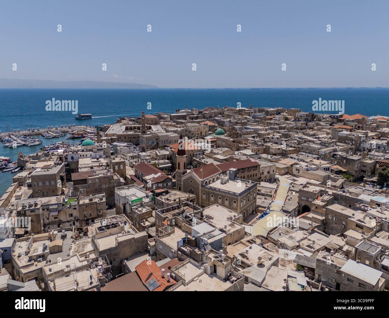 Vista aerea di una vivace città costiera con una guglia da chiesa dal tetto rosso che si innalza sopra la densa architettura, incontrando il tranquillo orizzonte blu del mare, acri, quartiere Nord, Israele. Foto Stock
