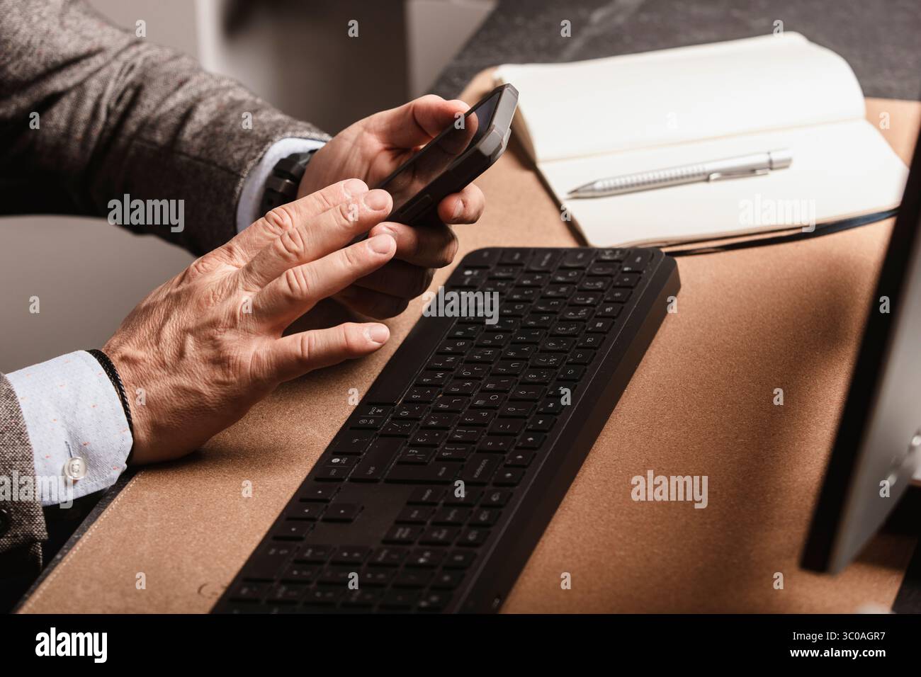 Uomo che utilizza lo smartphone alla moderna scrivania con tastiera, mouse, monitor e un bicchiere d'acqua. Area di lavoro minimalista, perfetta per lavorare in remoto Foto Stock