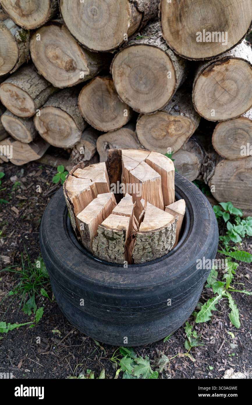 Legna da ardere divisa verticalmente impilata all'interno di un vecchio pneumatico con un pelo di legno di fondo: Concetto di riscaldamento rurale, trame di legno, soluzione ecologica, vita in campagna Foto Stock