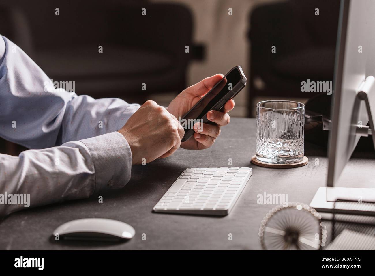 Uomo che utilizza lo smartphone alla moderna scrivania con tastiera, mouse, monitor e un bicchiere d'acqua. Area di lavoro minimalista, perfetta per lavorare in remoto Foto Stock