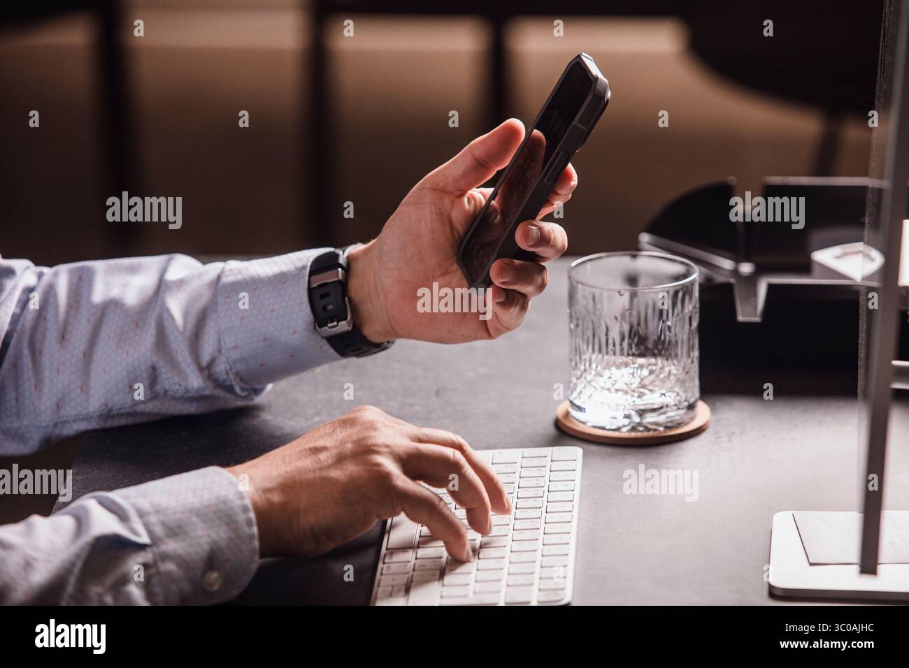 Uomo che utilizza lo smartphone alla moderna scrivania con tastiera, mouse, monitor e un bicchiere d'acqua. Area di lavoro minimalista, perfetta per lavorare in remoto Foto Stock