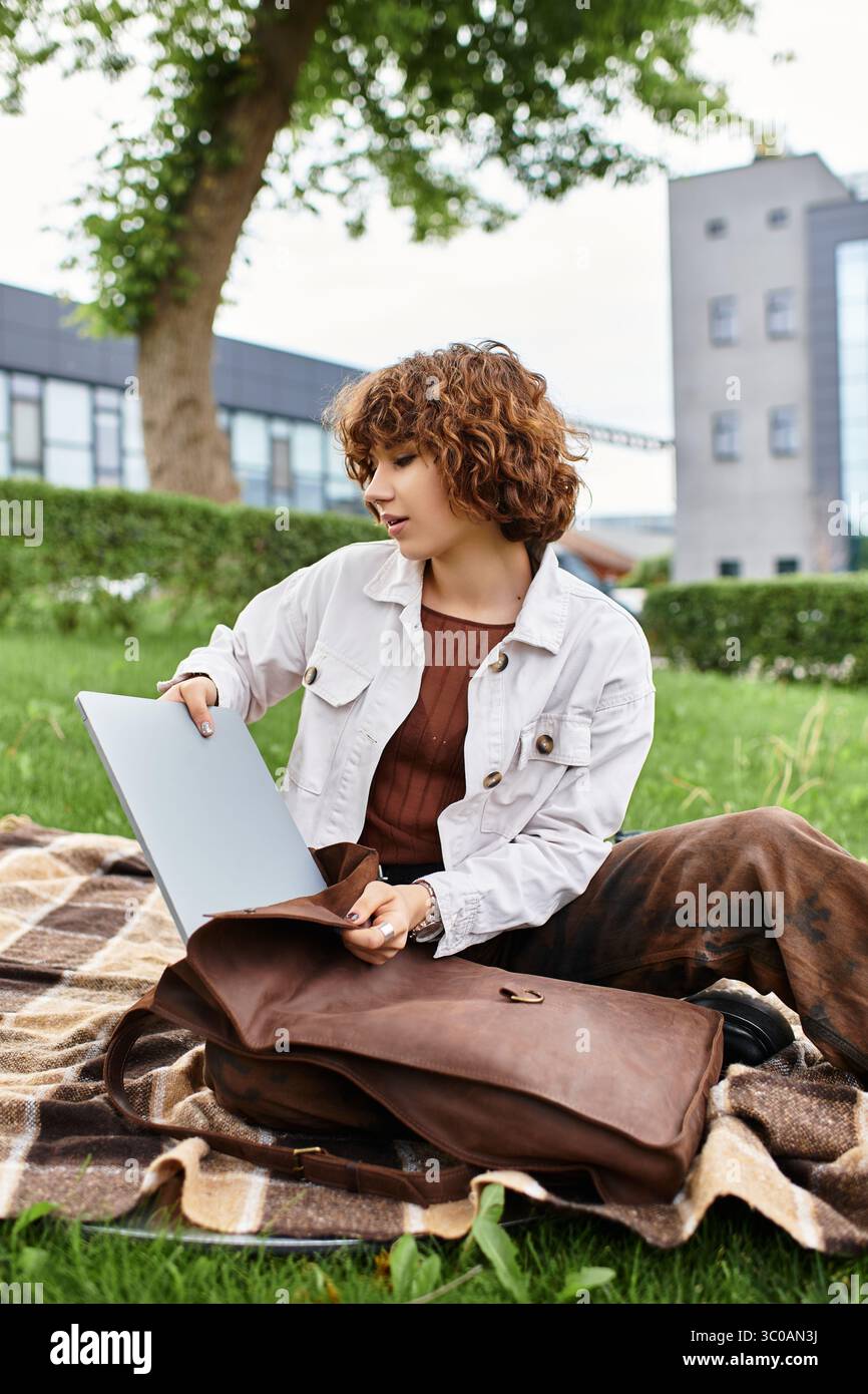 La giovane donna con i capelli ricci lavora in remoto sul suo notebook in un parco soleggiato. Foto Stock