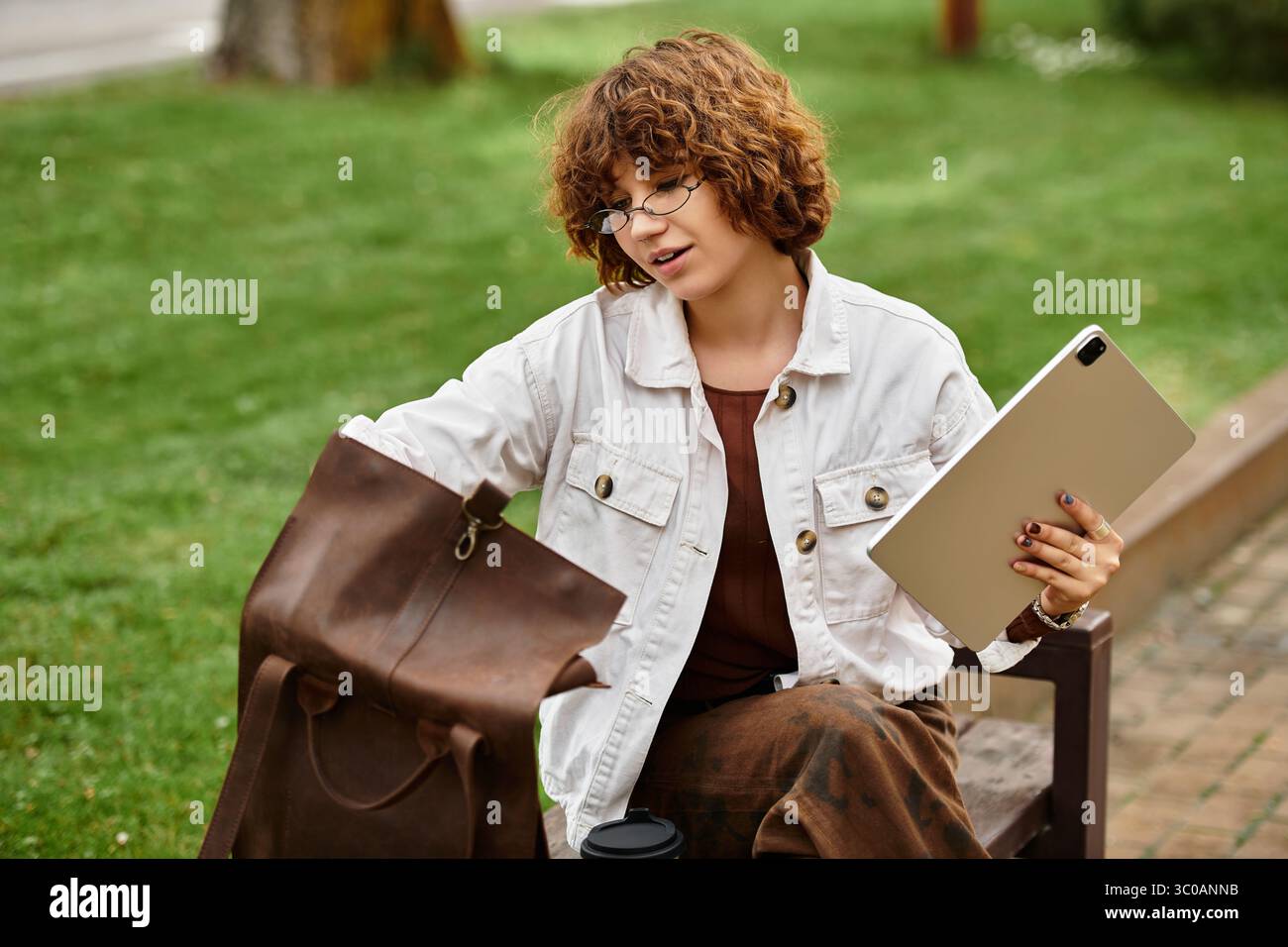 Una donna dai capelli ricci lavora in remoto in un parco soleggiato, bilanciando laptop e caffè. Foto Stock
