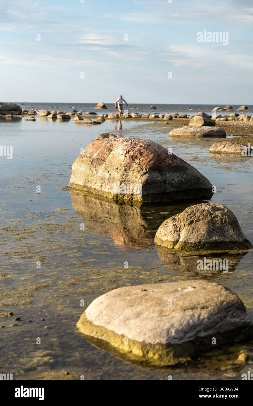 Figura solitaria che cammina tra le grandi rocce costiere di Käsmu, Estonia, una tranquilla scena costiera baltica con riflessi, natura, silenzio / presenza umana Foto Stock
