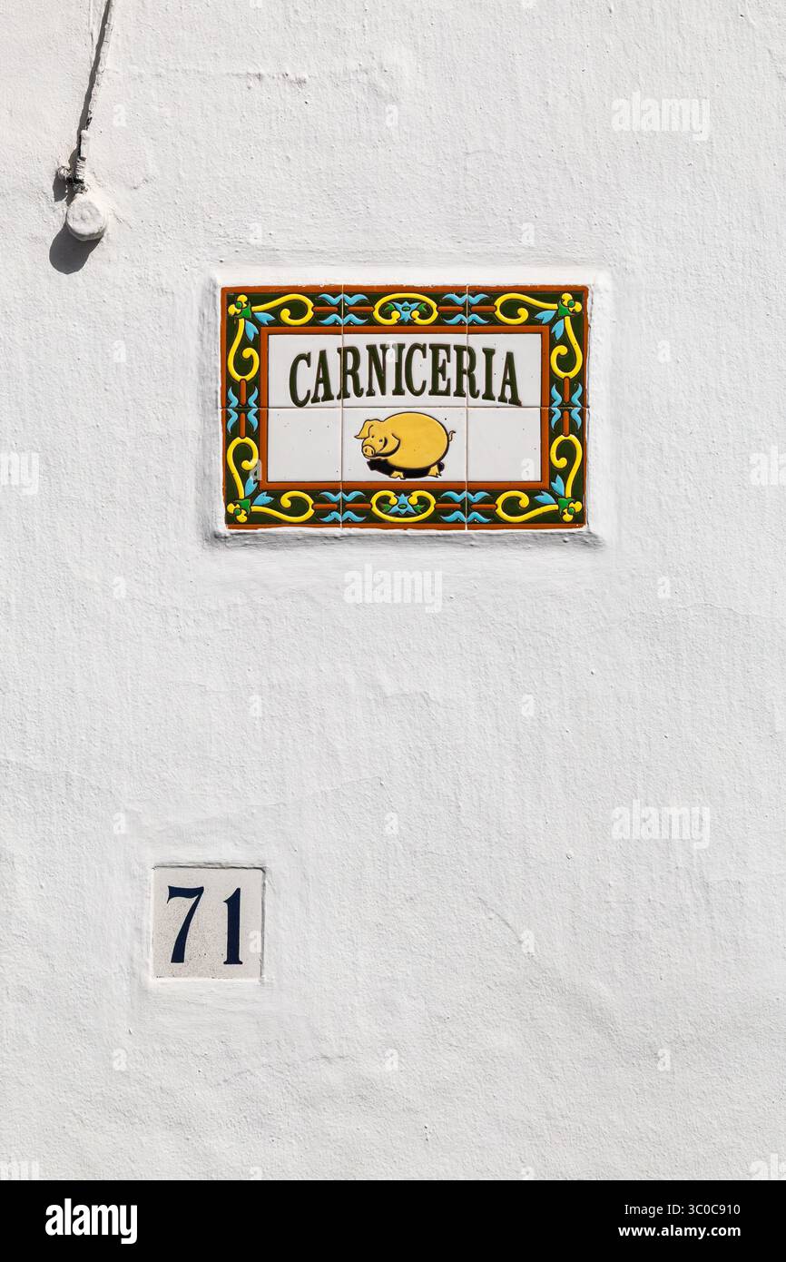 Insegna di piastrelle in ceramica colorata con un'illustrazione di maiale contrassegnata con Carniceria (macelleria) su una parete bianca accanto a un cancello in metallo decorativo. Frigiliana, Spagna Foto Stock
