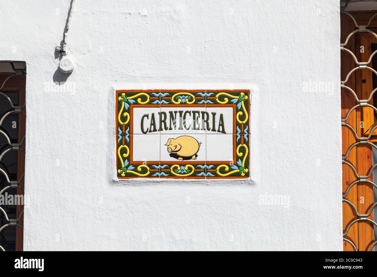 Insegna di piastrelle in ceramica colorata con un'illustrazione di maiale contrassegnata con Carniceria (macelleria) su una parete bianca accanto a un cancello in metallo decorativo. Frigiliana, Spagna Foto Stock