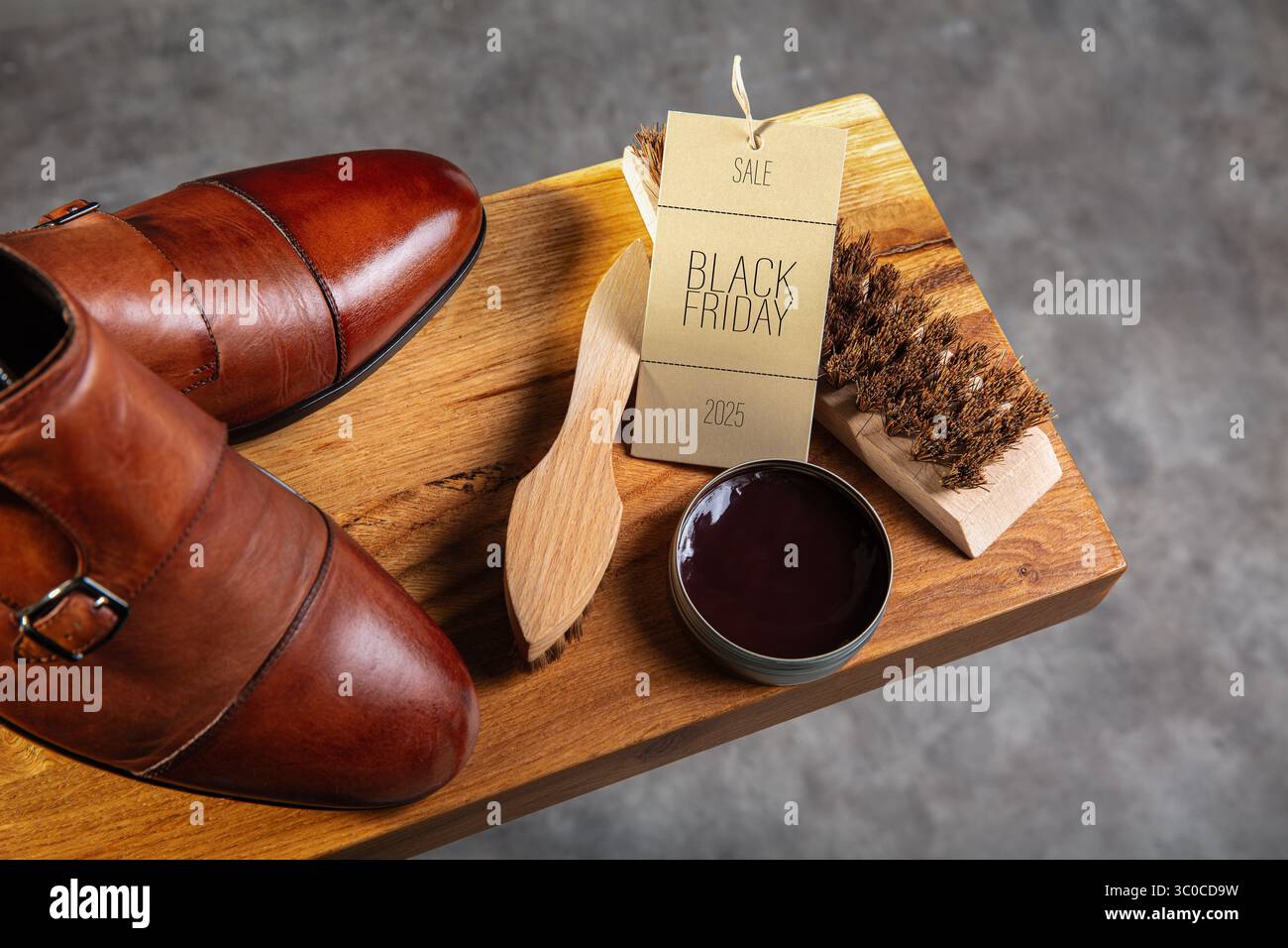 "Vista dall'alto delle scarpe con cinturino in pelle marrone lucida posizionate accanto a una scatola di lucidatura per scarpe, spazzole in legno e un cartellino di vendita "Black Friday 2025"." Il compo Foto Stock