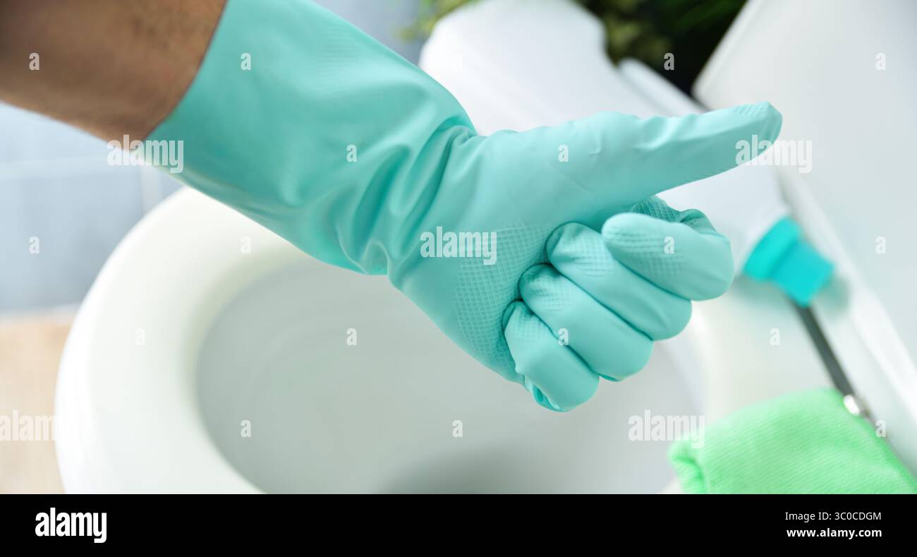 Mano con guanto di gomma blu scrubbing con gesti ok e sfondo con ciotola igienica appena disinfettata con coperchio aperto in un bagno blu. Via sopraelevata Foto Stock