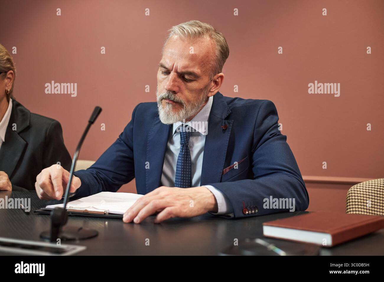 Uomo caucasico di mezza età seduto al tavolo da conferenza scrivendo su documento con penna, vestito e cravatta, partecipando a un incontro politico con microfono e libro visibile Foto Stock