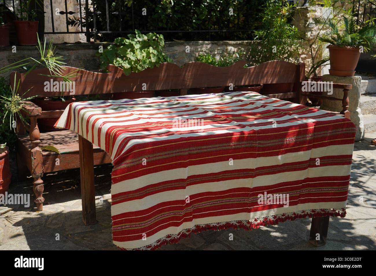 Tradizionale panca e tavolo in legno ricoperti da un tessuto a righe rosso e bianco, presso il cortile del Monastero di Kallergi. Atmosfera estiva soleggiata. Foto Stock