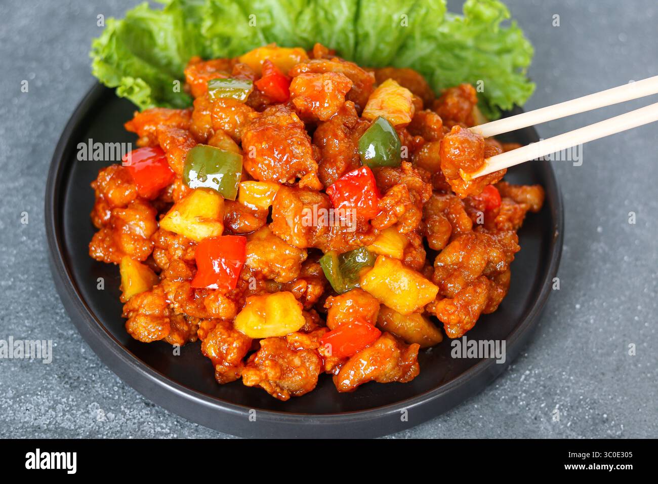 Pollo agrodolce servito su un piatto scuro, con un paio di bacchette che ne raccolgono un pezzo. Decorata con peperoni e pezzi di ananas. Foto Stock