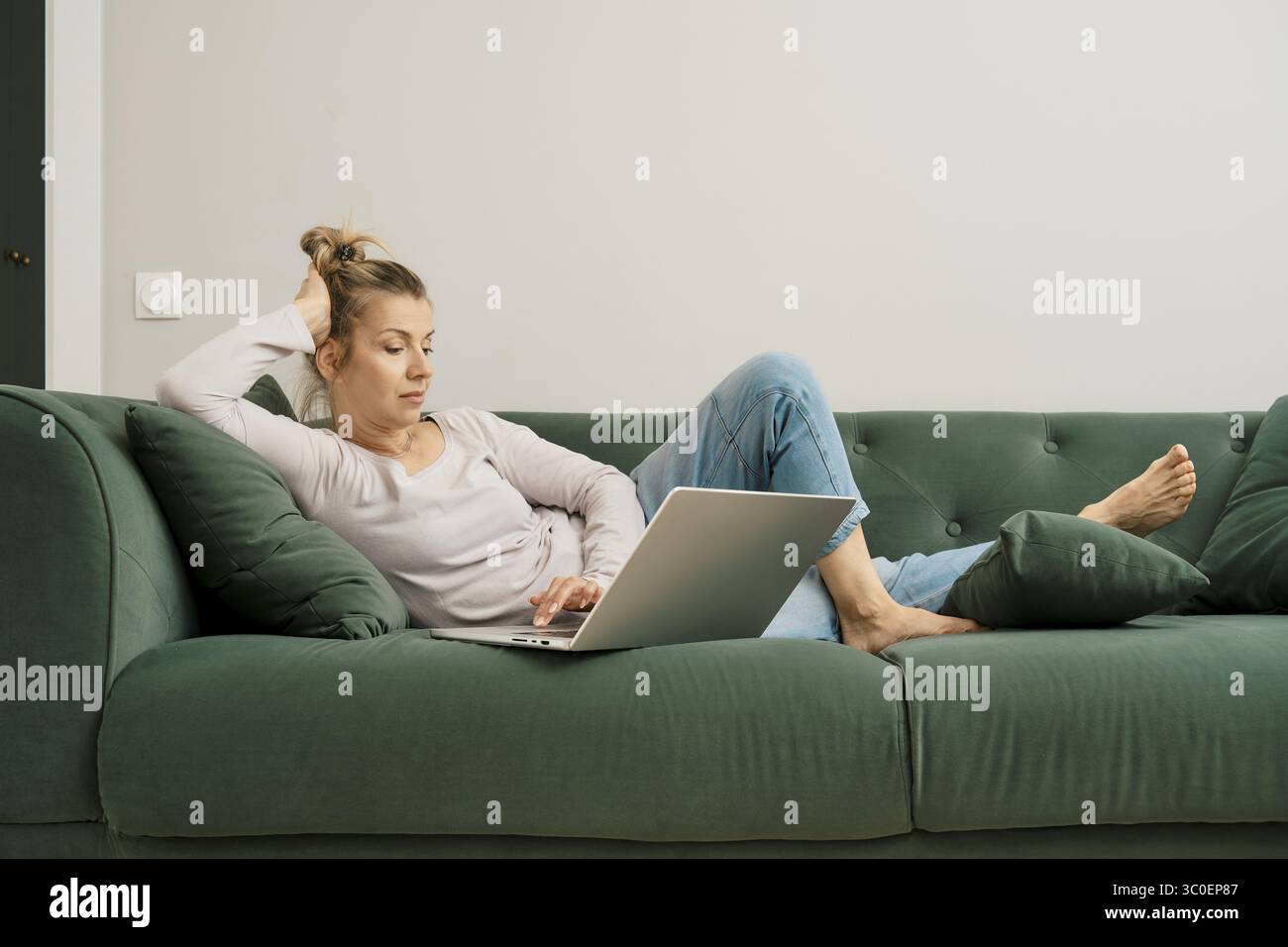 Una donna è seduta su un divano verde, scorrendo i social media sul suo laptop. Indossa abiti casual e i capelli vengono tirati indietro Foto Stock