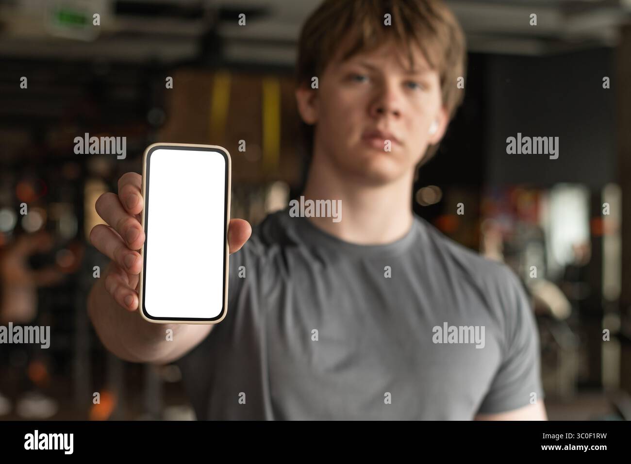 Atleta in piedi e che mostra il cellulare con schermo bianco bianco in un ambiente da palestra sfocato. App fitness. Immagine di un mockup con un uomo sportivo. Spazio per il design. Foto Stock