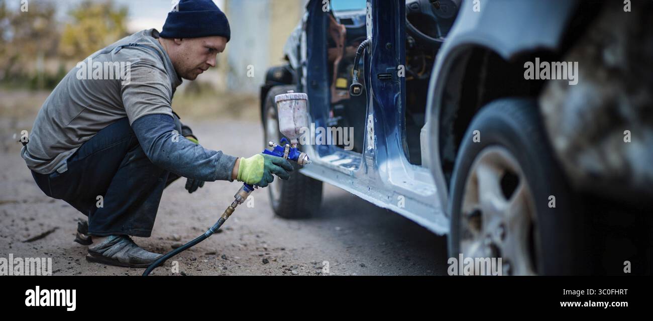 Un uomo che accovacciava, dipingeva un'auto con una pistola a spruzzo all'aperto Foto Stock