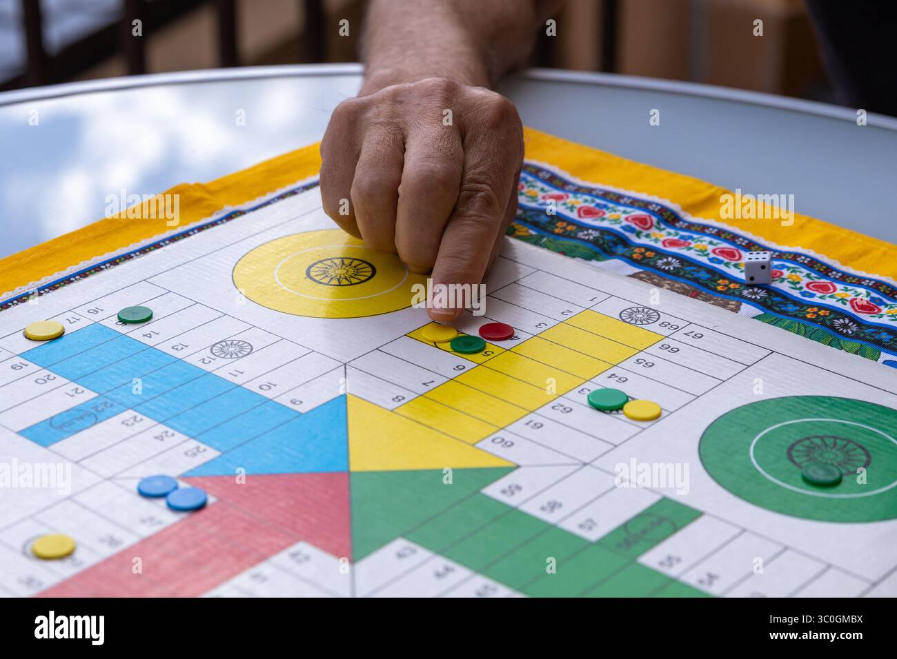 Mano di un pensionato che muove una pedina rossa su una tavola parcheesi colorata, godendosi un classico gioco da tavolo durante il tempo libero Foto Stock