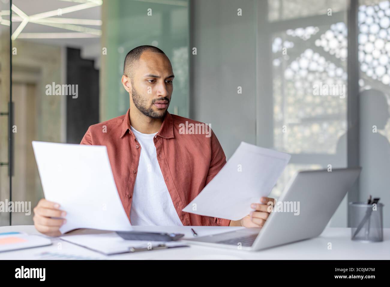 Un uomo d'affari concentrato che esamina i documenti cartacei, studia documenti in un ufficio moderno, accanto a un notebook e ad altri oggetti correlati al lavoro. Foto Stock