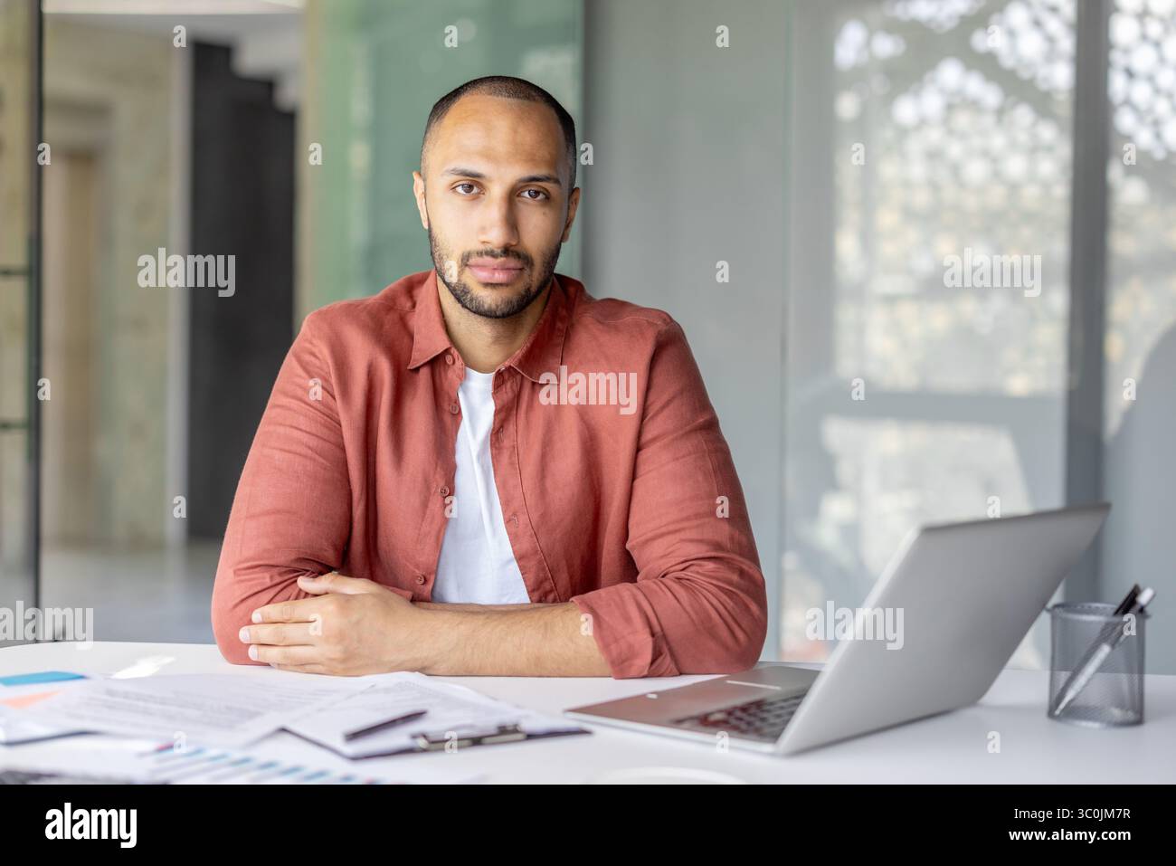 Un uomo premuroso si siede a una scrivania, guardando direttamente la fotocamera, con un portatile e documenti nelle vicinanze. Foto Stock