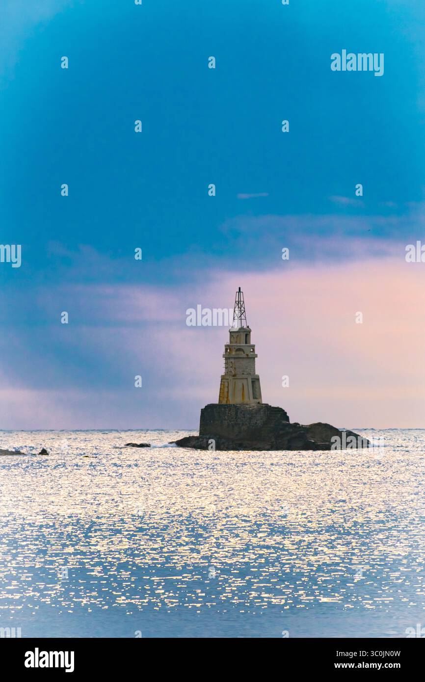 Un faro arroccato su un'isola rocciosa si erge alto in acque tranquille al crepuscolo, circondato da dolci onde e un vasto cielo dipinto con sottile h Foto Stock