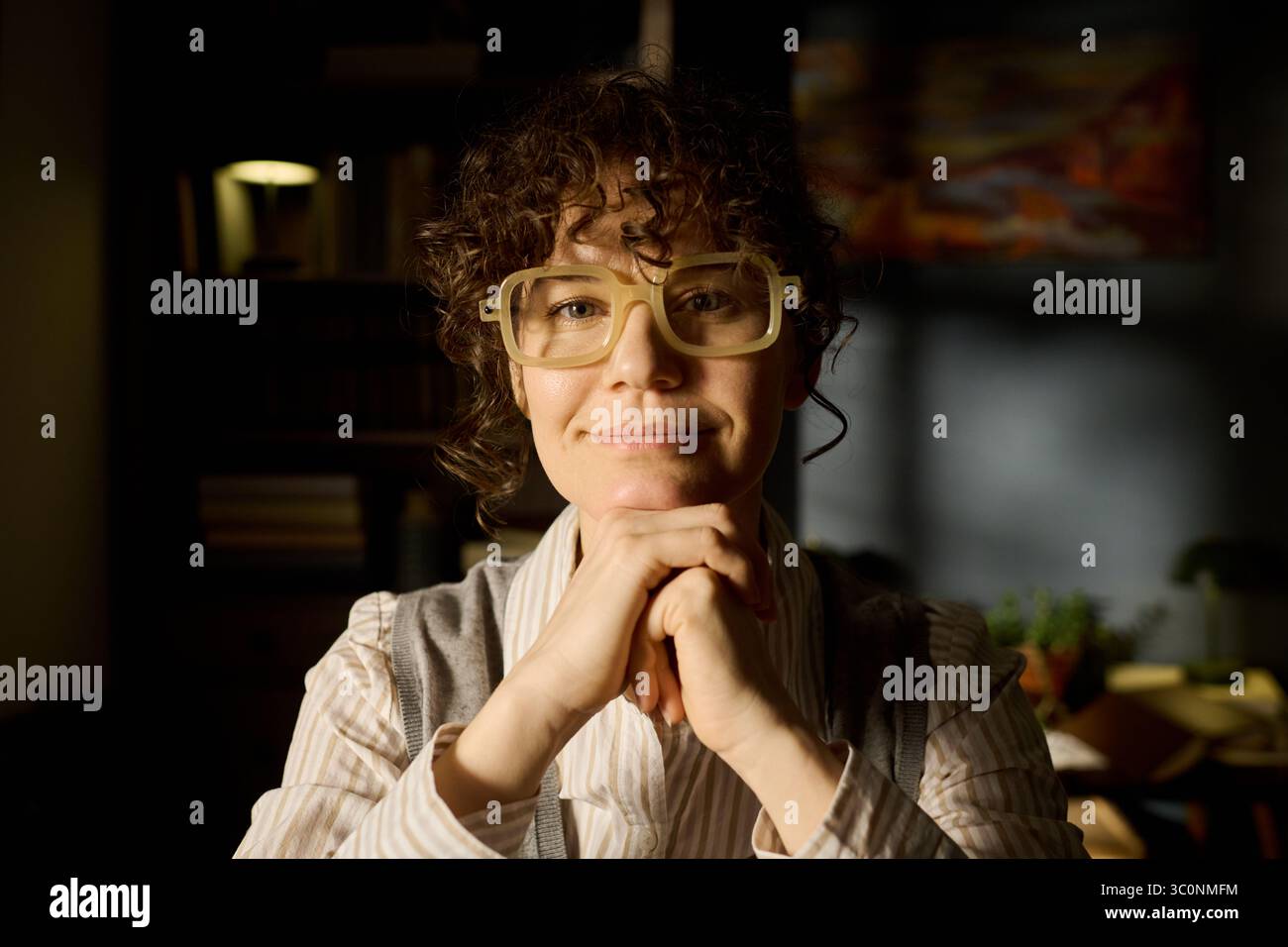 Ritratto di una donna caucasica di mezza età con capelli ricci e occhiali che sorridono delicatamente mentre si riposa il mento sulle mani, guardando direttamente la fotocamera in un ambiente interno con luce soffusa Foto Stock