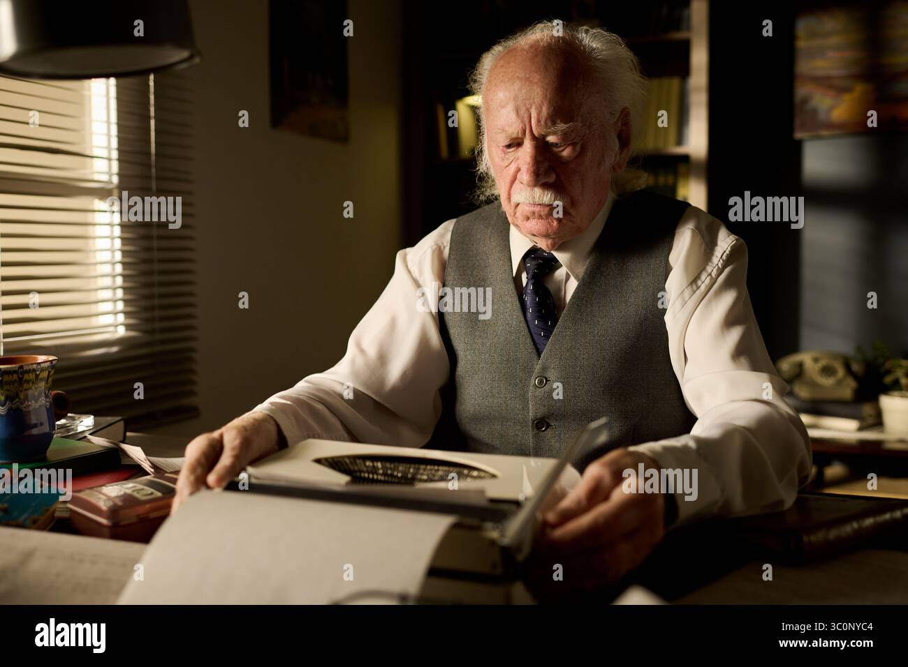 Uomo caucasico seduto alla scrivania usando una macchina da scrivere, concentrandosi sulla scrittura di documenti, indossando camicia e gilet formali, circondato da libri e documenti in ufficio Foto Stock