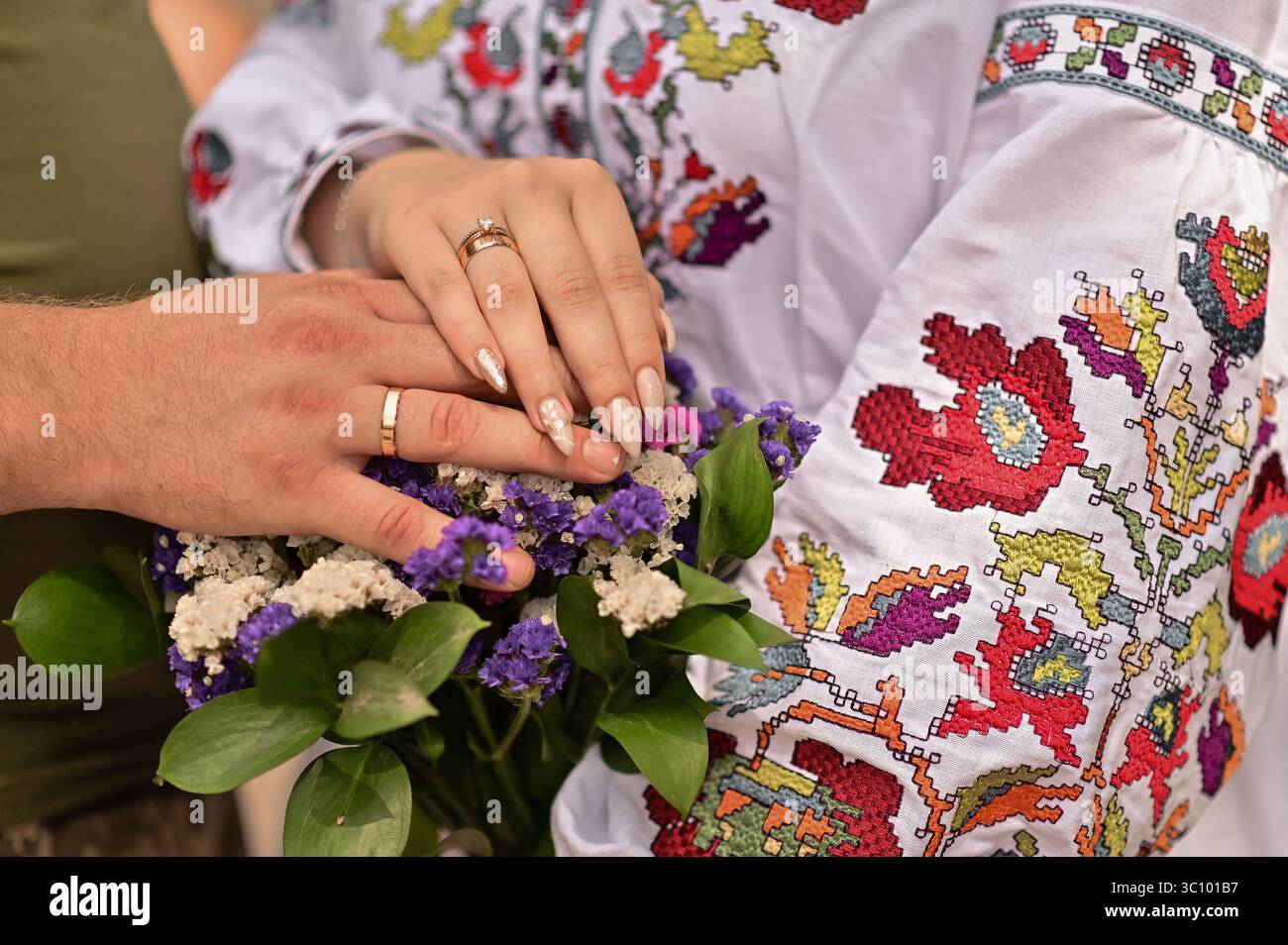 Primo piano delle mani di coppia con le fedi nuziali, con un bouquet, vestito con le tradizionali camicie ricamate ucraine. Momento romantico che simboleggia la lov Foto Stock