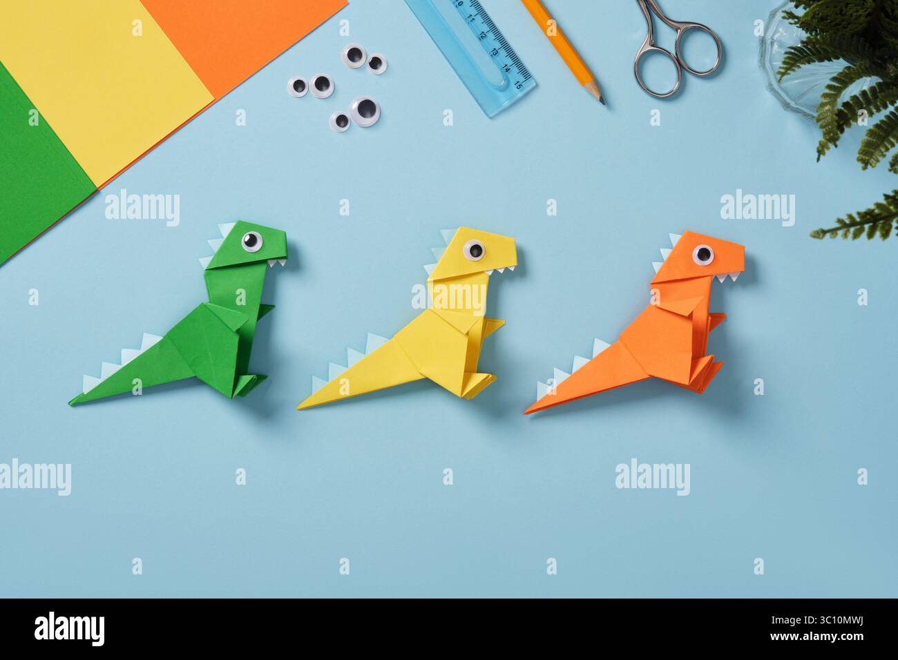Tavolo da lavoro Origami con fogli di carta, occhielli, forbici, flacone di colla, righello, e gomma su sfondo azzurro. Dinosauro di carta finito Foto Stock