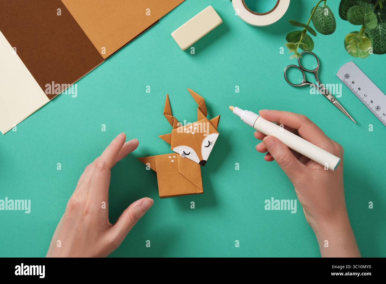 Le mani disegnano punti bianchi con un pennarello su un'imbarcazione di carta delle renne. Intorno a loro ci sono rifornimenti di carta artigianale. Foto Stock