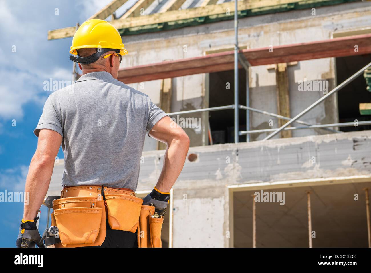 L'operaio edile con un casco giallo ispeziona il cantiere. Ponteggi e pareti non finite sono visibili, mostrando i lavori in corso durante il giorno Foto Stock