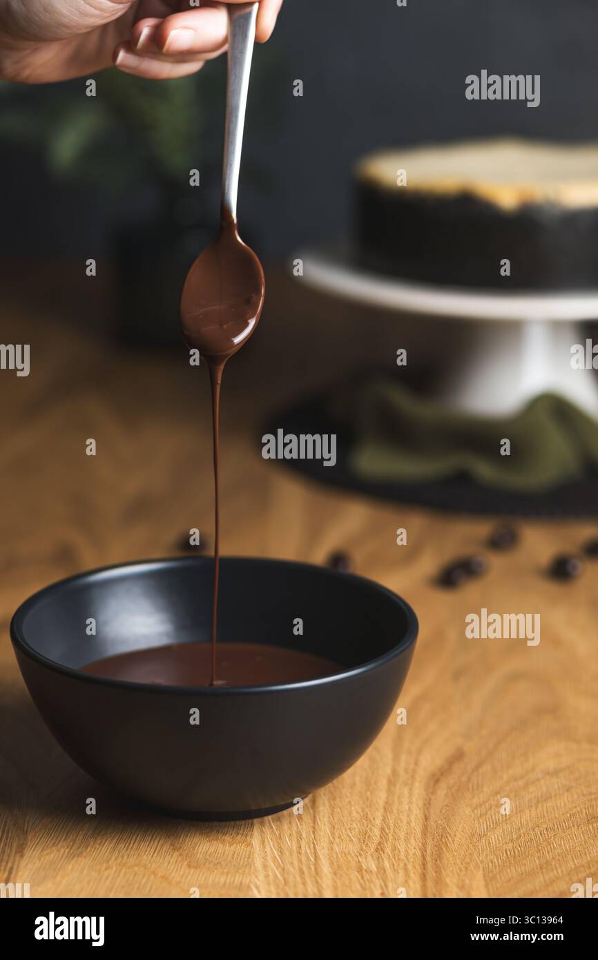 Tenere a mano un cucchiaio che gocciola il cioccolato liscio fuso in una ciotola nera, con una cheesecake sullo sfondo. Foto Stock