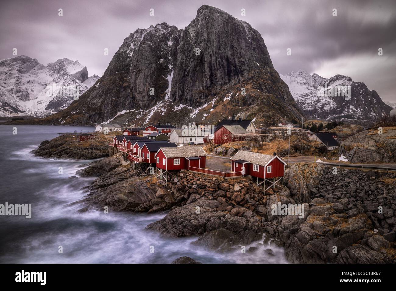 Veduta delle impressionanti cabine rosse che si aggrappano alle rocce scoscese dalle onde sotto montagne colossali e innevate sotto un cielo moodo, Hamnøy, Norvegia. Foto Stock