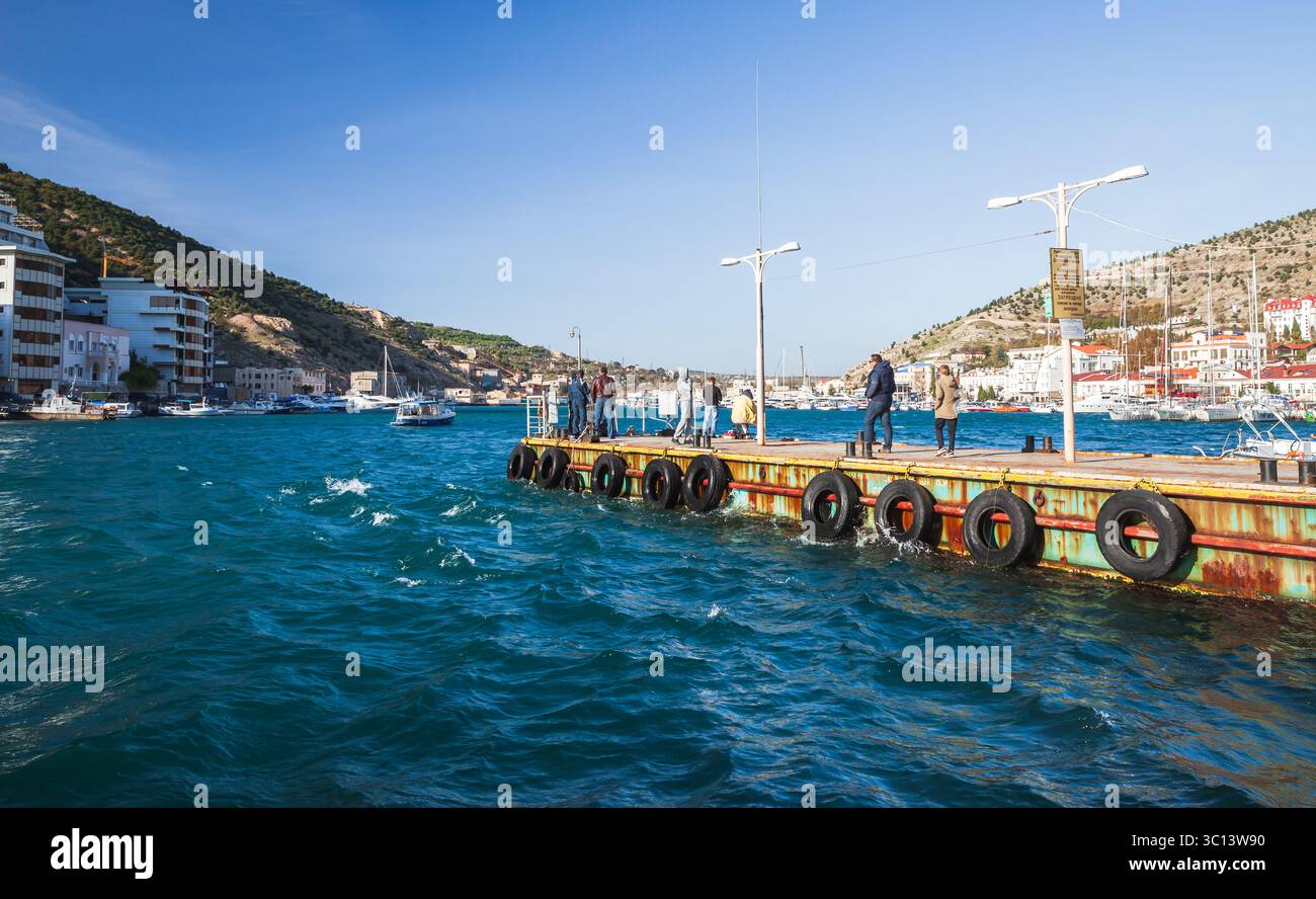 Balaklava, Crimea - 28 ottobre 2018: Persone che si godono la vista da un molo panoramico con una città costiera e barche sullo sfondo Foto Stock