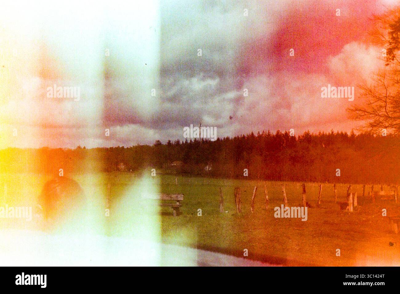 Paesaggio analogico con spettacolari perdite di luce, che mostra un campo erboso, recinzioni in legno e una foresta con una torre radio. Girato su pellicola da 35 mm, Paesi Bassi. Foto Stock