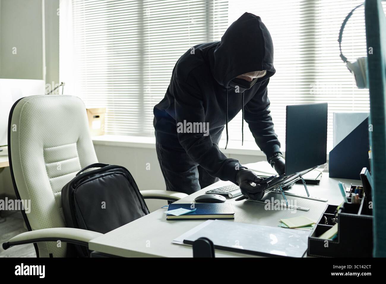 Uomo anonimo con cappuccio. maschera nera e guanti che rubano il laptop dalla moderna scrivania dell'ufficio, in piedi accanto allo zaino aperto e guardando verso il basso il dispositivo, persiane che coprono la finestra sullo sfondo Foto Stock