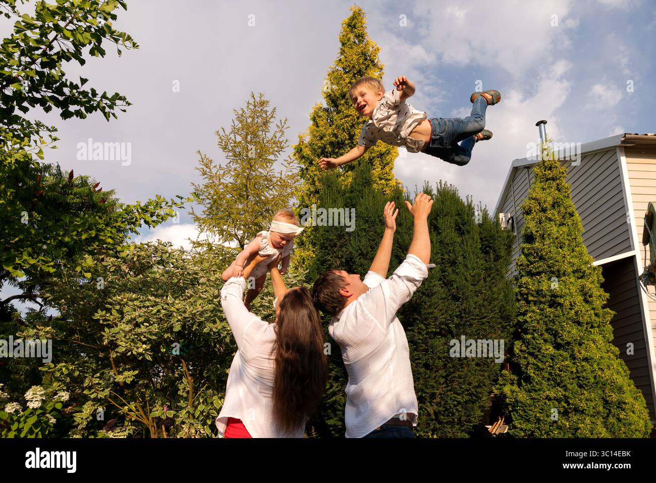 Nella luce della sera dorata, il padre getta in alto il figlio ridendo, mentre la madre solleva il bambino, giocherellando in famiglia incorniciato da alti sempreverdi e backya Foto Stock