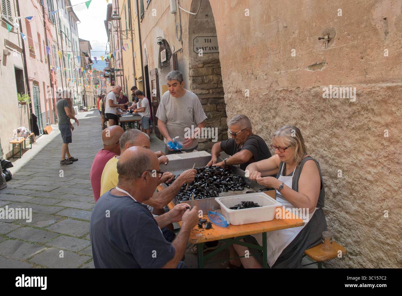 Festival gastronomici in Italia. L'annuale festa dei molluschi di cozze Borgomaro nel borgo medievale italiano. La gente della strada principale, che apre i molluschi, prepara il pasto per la prossima sera. Borgomaro, regione Liguria, Italia, Europa. Luglio 2025 HOMER SYKES Foto Stock