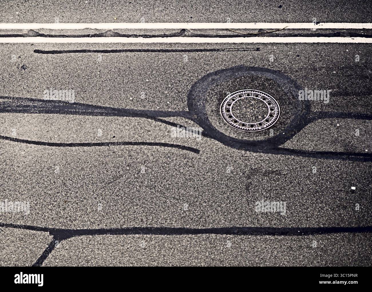 Una copertura rotonda per tombini e linee stradali formano un modello geometrico su una superficie asfaltata ruvida, Reifenspuren Asphalt Streetview Foto Stock