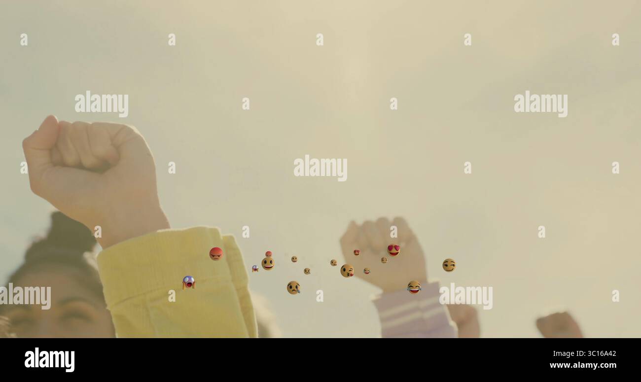 Alzare i pugni donne che indossano maniche colorate che si uniscono in spazi esterni con emoji a reazione fluttuante Foto Stock
