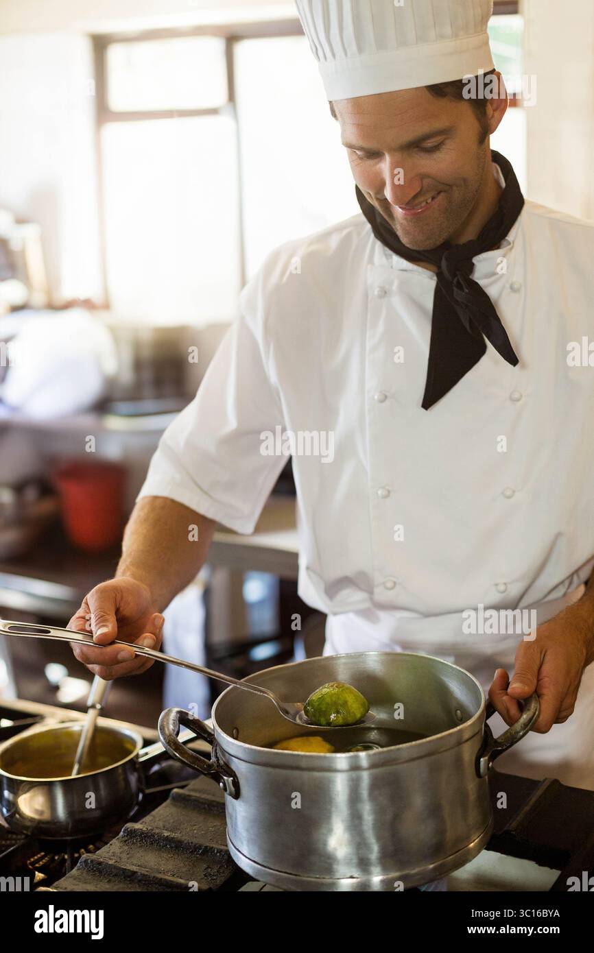 Lo chef maschile indossa un'uniforme che mescola verdure verdi in pentola di acciaio su una stufa a gas nella cucina del ristorante Foto Stock
