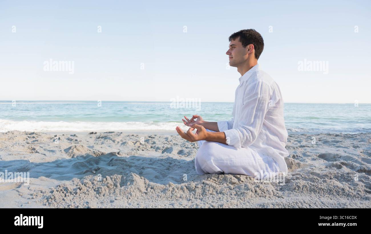 Uomo seduto a gambe incrociate a meditare sulla costa sabbiosa di fronte all'oceano sotto il cielo limpido, copia lo spazio Foto Stock