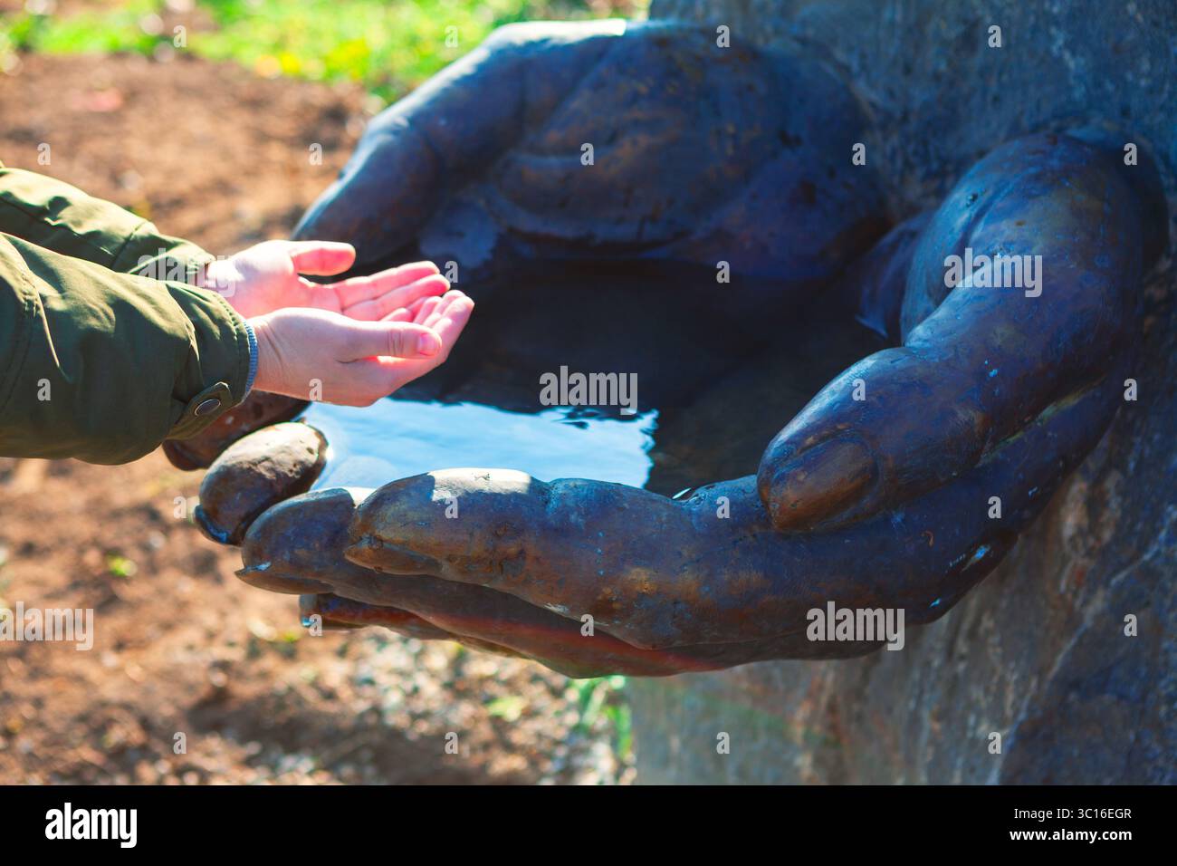 Persona con le mani allungate che si avvicina alla scultura in pietra più grande di mani chiuse che reggono l'acqua. La scena è ambientata all'aperto con uno sfondo naturale Foto Stock