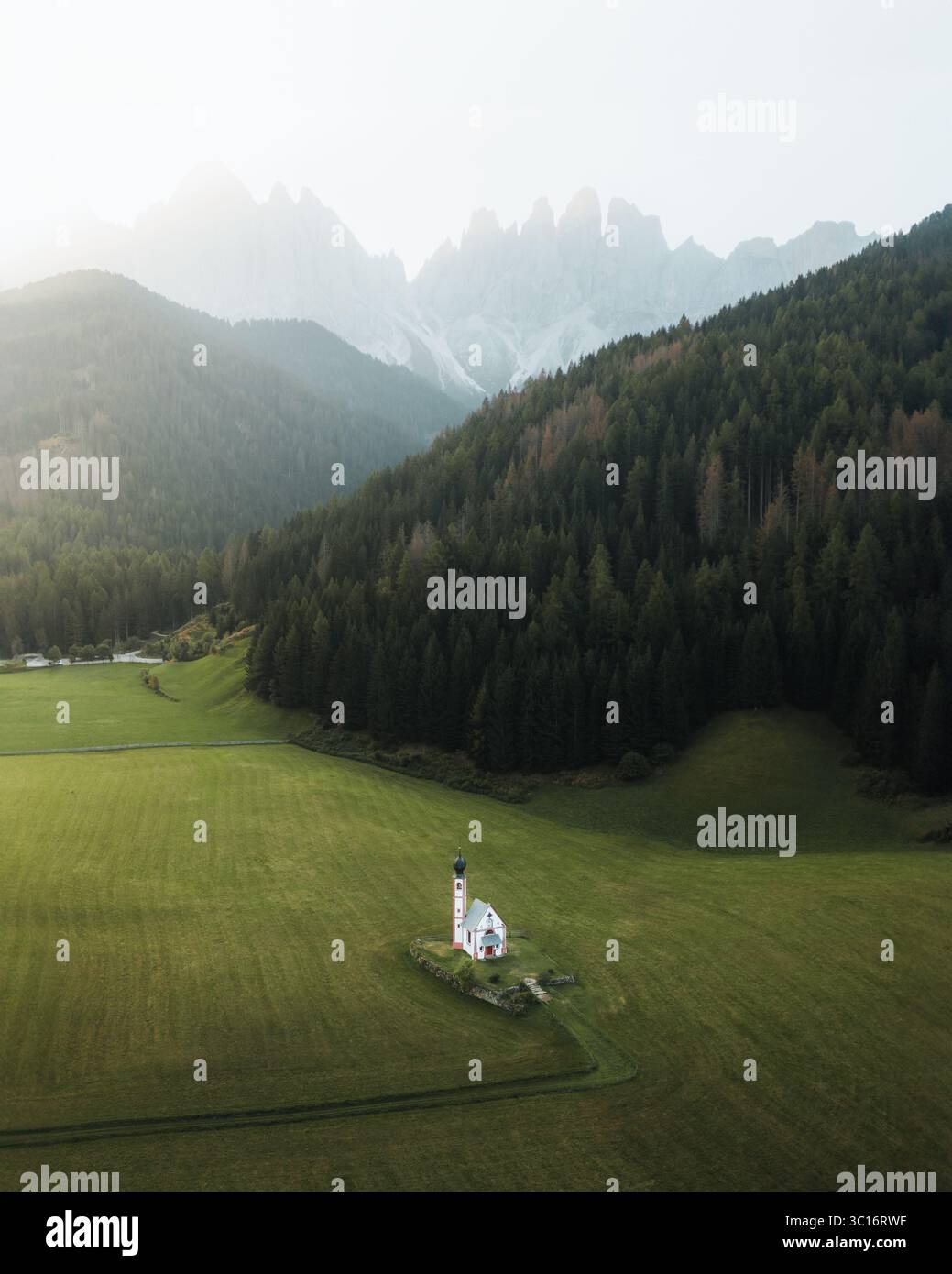 Veduta aerea di una chiesa solitaria immersa in un campo verde vivido, abbracciata da una fitta foresta e montagne torreggianti, Val di Funes, Trentino-alto A. Foto Stock