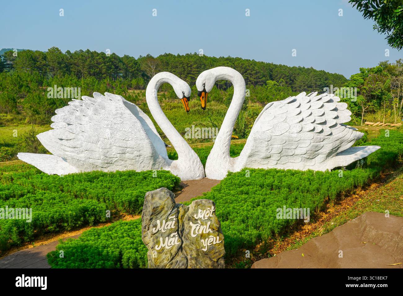 Da Lat, Vietnam - 24 marzo 2025: Due maestose sculture di cigno bianco formano la forma del cuore con il collo, simboleggiando l'amore eterno in mezzo a una vegetazione lussureggiante Foto Stock