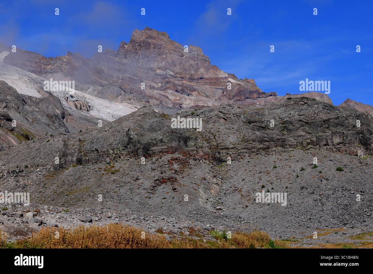 Le scogliere vulcaniche illuminate lateralmente e i campi innevati si innalzano sopra i pendii nebbiosi del talco sotto un cielo blu profondo, rivelando i dettagli delle aspre strutture alpine del Monte Rainier. Foto Stock