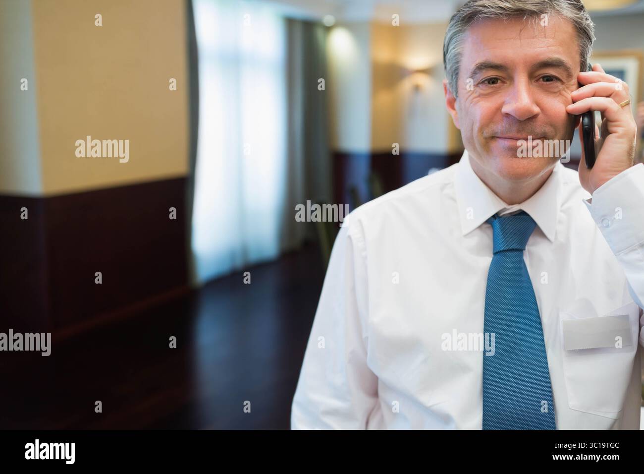 Uomo maturo in piedi nel corridoio dell'hotel con smartphone e badge per conferenze, spazio per fotocopie Foto Stock