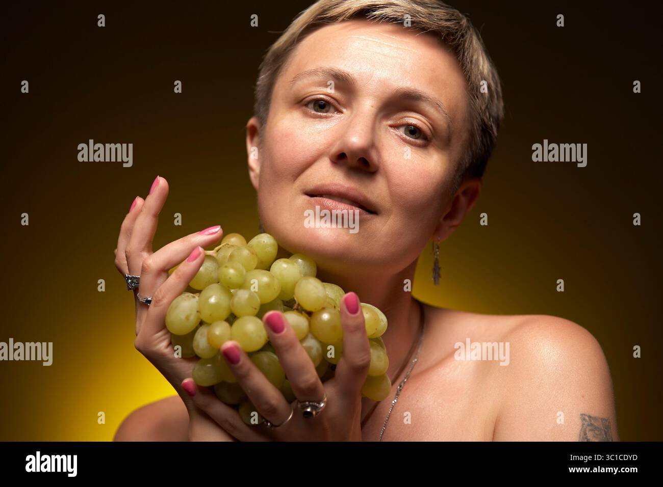 Una donna con i capelli biondi corti tiene in mano un mucchio di uva verde. Ha un sorriso sottile. Indossa una collana d'argento e uno smalto rosa. T Foto Stock