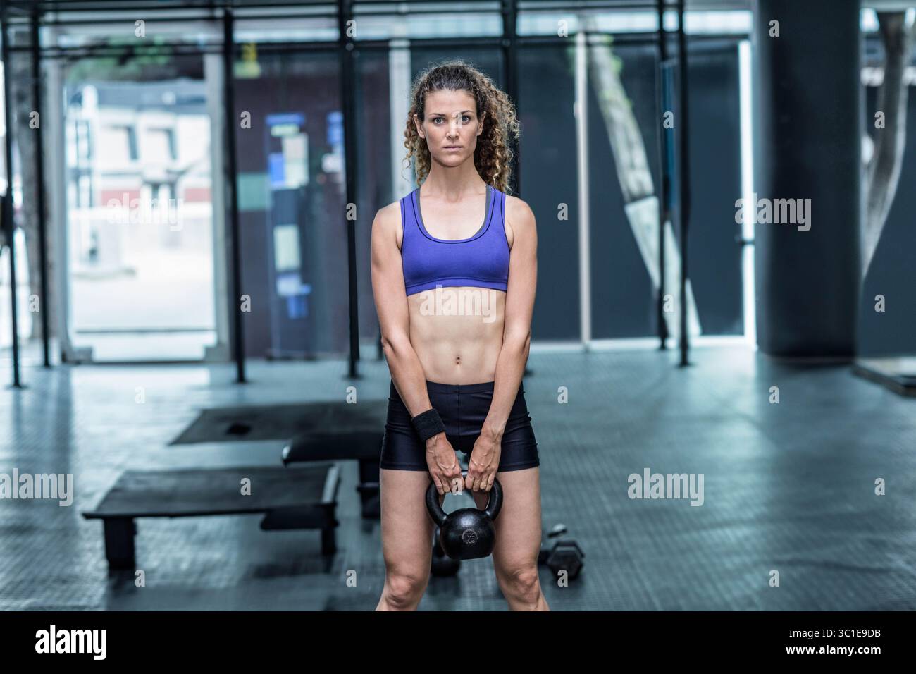 Allenatrice di abbigliamento sportivo che solleva il campanello nero all'interno della palestra accanto alla panca per pesi, manubrio Foto Stock