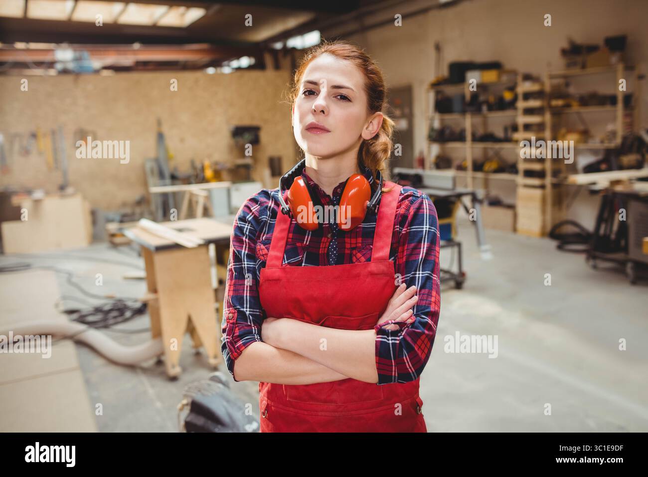 Lavoratrice del legno che indossa grembiule e cuffie rosse, in piedi tra banchi da lavoro e legname in officina Foto Stock