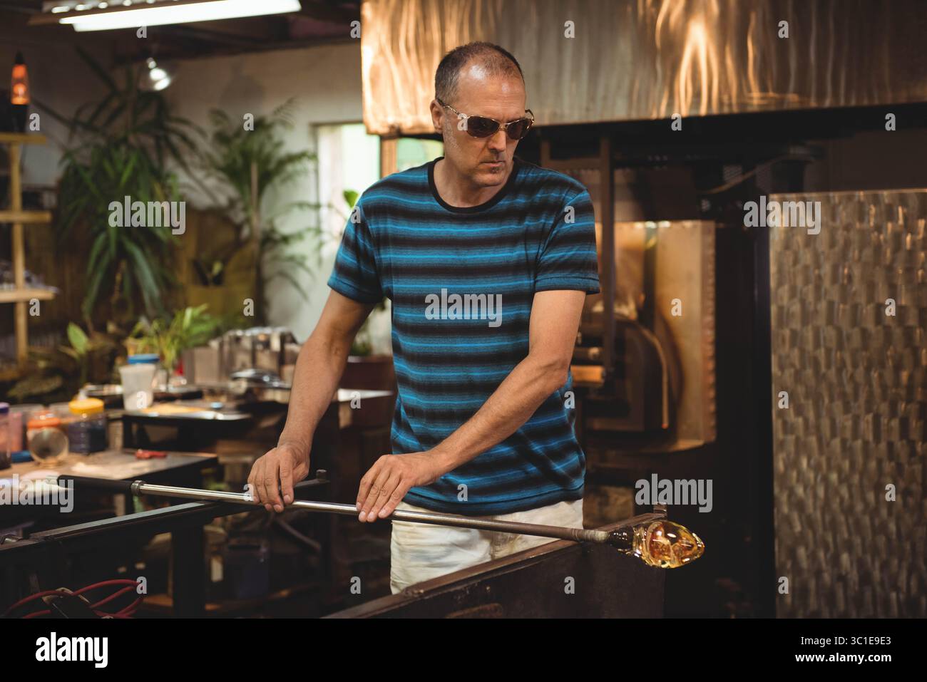 Uomo di mezza età in piedi sul banco di soffiatura del vetro afferrando il tubo di soffiaggio con vetro fuso nella fornace Foto Stock